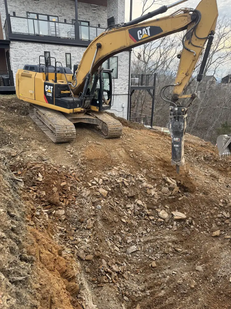 Excavator with hammer attachment breaking rock on a hillside site